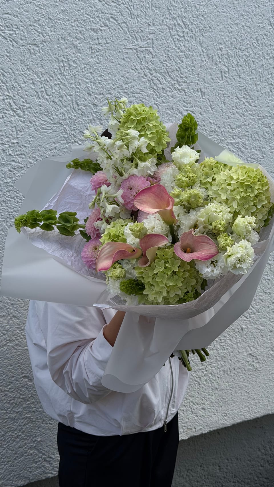 Lockerer Strauß aus weißen und grünen Blüten mit rosa Callas vor einer weißen Wand