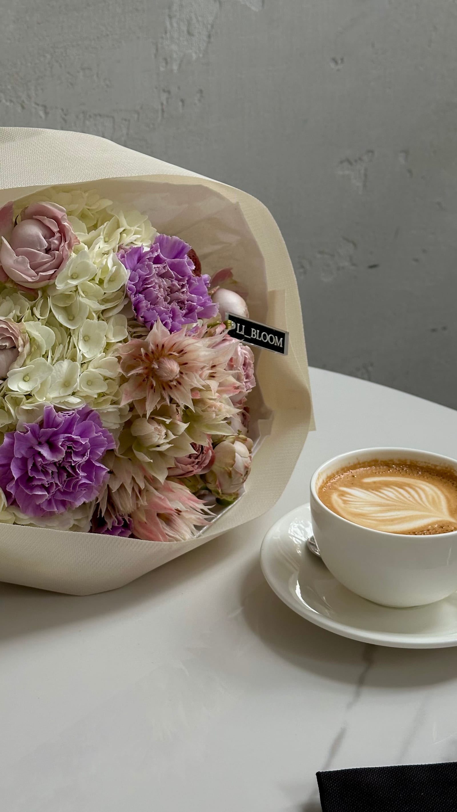 Strauß in Flieder und Altrosa auf einem weißen Tisch, daneben eine Tasse Kaffee