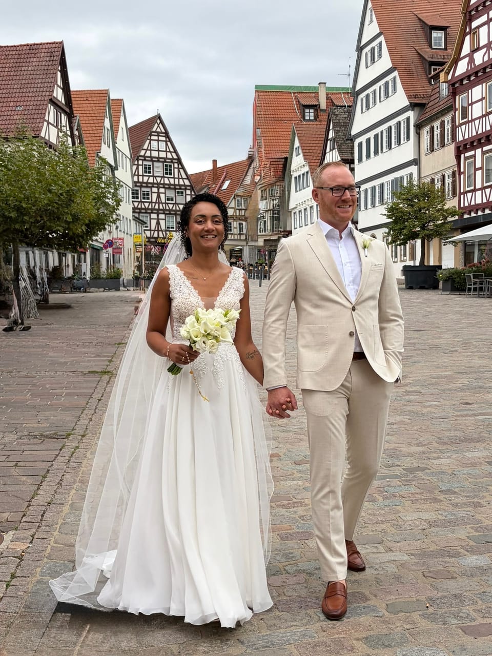 Brautpaar Hand in Hand auf einer gepflasterten Altstadtgasse mit Fachwerkhäusern, die Braut mit Schleier hält einen Strauß aus weißen Callas