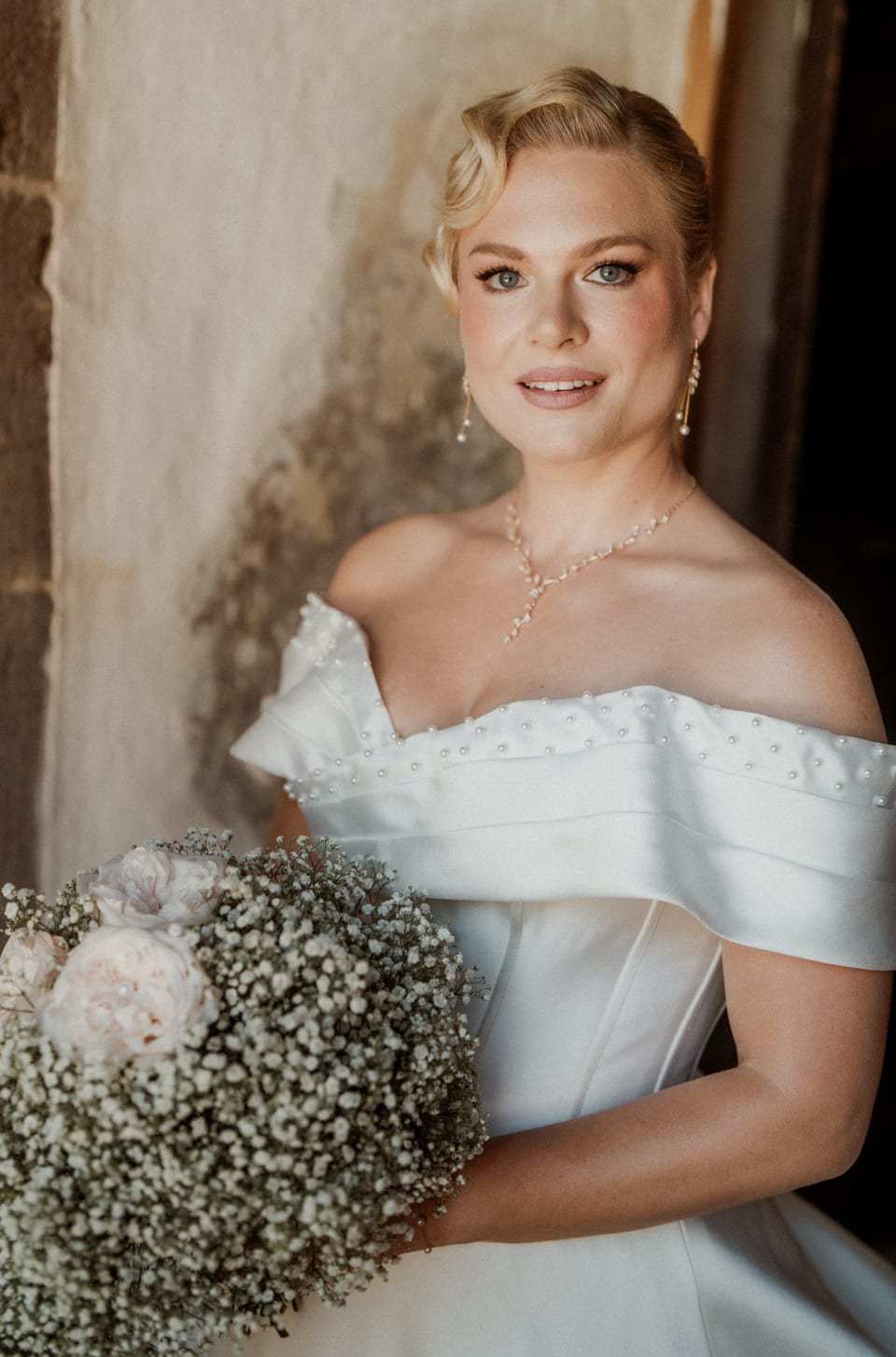 Braut mit Wasserwellen im schulterfreien Kleid mit Perlen, im Arm ein Strauß aus Schleierkraut und zartrosa Rosen